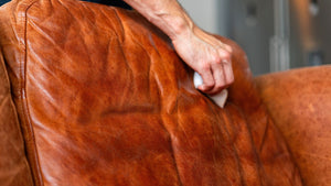 A close-up of a person's hand using a white cloth to clean the creases of a warm brown leather armchair.