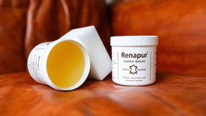 A jar of Renapur Leather Balsam shown next to an open white container revealing the yellow-colored balm inside, set on a brown leather surface.