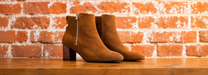 Tan suede ankle boots on a wooden surface against a brick wall.