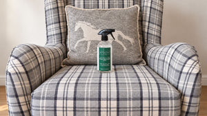 A bottle of Renapur Suede, Nubuck & Fabric Protecter placed on the seat of a plaid armchair, with a matching plaid armchair and a gray throw pillow featuring a white horse silhouette in the background.