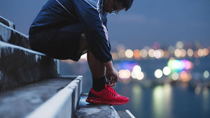 A person crouching on outdoor concrete steps at dusk, tying the shoe of a bright red athletic sneaker. The background shows a blurred city waterfront with colorful bokeh lights.