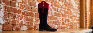 Blue suede boot with a red cuff on a wooden shelf against a brick wall.