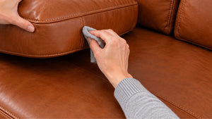 Testing leather cleaner with a soft grey cloth on the side of a lifted brown leather sofa cushion.