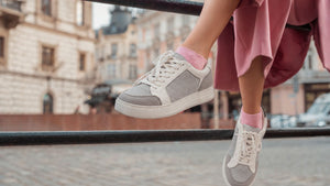 A person wearing light gray and white low-top sneakers with pink socks, sitting on a black metal railing in an outdoor urban setting with blurred buildings in the background.