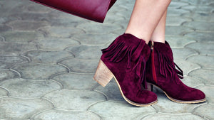A close-up of a person wearing burgundy suede ankle boots with fringe detailing and block heels, standing on a gray paved surface.
