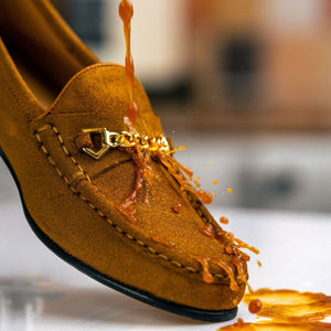 Tea pouring over a tan suede loafer with gold chain detailing and running off the toe.